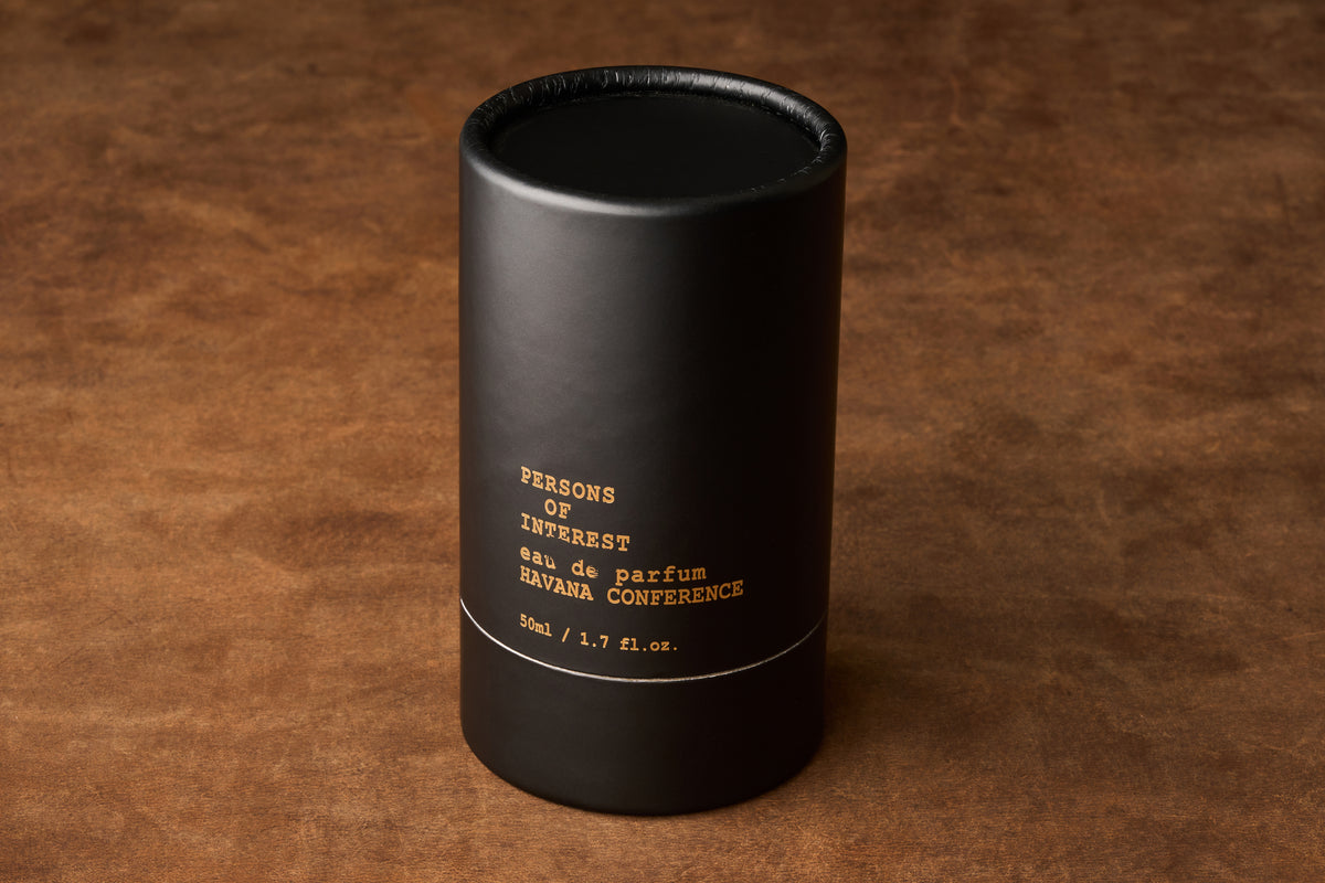A black cylindrical bottle with gold lettering displaying “Persons of Interest Havana Conference Eau de Parfum 50ml / 1.7 fl. oz.” rests on a brown textured surface, evoking the sophisticated rum and subtle cedarwood notes of this refined fragrance.
