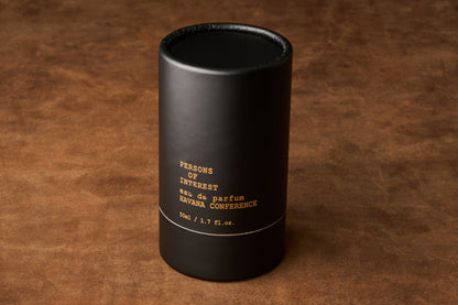 A black cylindrical bottle with gold lettering displaying “Persons of Interest Havana Conference Eau de Parfum 50ml / 1.7 fl. oz.” rests on a brown textured surface, evoking the sophisticated rum and subtle cedarwood notes of this refined fragrance.