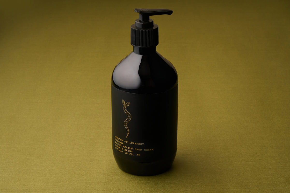 A black pump bottle of Persons of Interest Satsuma Active Relief Hand Cream, featuring gold text and a caduceus symbol, rests on an olive-green textured surface. This deeply hydrating hand cream is both sulfate- and paraben-free.