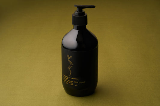 A black pump bottle of Persons of Interest Satsuma Active Relief Hand Cream, featuring gold text and a caduceus symbol, rests on an olive-green textured surface. This deeply hydrating hand cream is both sulfate- and paraben-free.