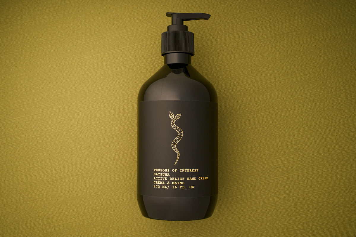 A black lotion bottle with a pump sits on an olive green textured surface. The label features a gold snake design and gold text describing the product: Persons of Interest Satsuma Active Relief Hand Cream, a deeply hydrating hand cream that is sulfate and paraben free.