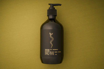 A black lotion bottle with a pump sits on an olive green textured surface. The label features a gold snake design and gold text describing the product: Persons of Interest Satsuma Active Relief Hand Cream, a deeply hydrating hand cream that is sulfate and paraben free.