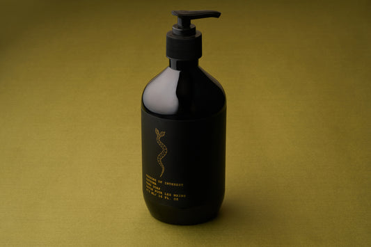 A black pump bottle of Persons of Interest Satsuma Hand Soap, featuring minimal gold text and a snake design, stands on an olive green surface with a matching green background. This paraben-free soap offers a refreshing satsuma scent.