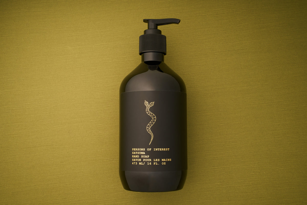 A dark bottle of Persons of Interest Satsuma Hand Soap with a pump rests on an olive green surface. The label features a gold snake design and gold text detailing the product information, highlighting its paraben-free formula and refreshing satsuma scent.