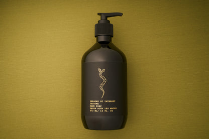 A dark bottle of Persons of Interest Satsuma Hand Soap with a pump rests on an olive green surface. The label features a gold snake design and gold text detailing the product information, highlighting its paraben-free formula and refreshing satsuma scent.