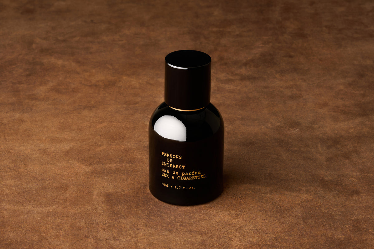 A black bottle of Persons of Interest Sex & Cigarettes Eau de Parfum (50ml / 1.7 fl. oz.) rests on a brown textured surface, alluding to its smoky fragrance with notes of tobacco, bergamot, and cedarwood.
