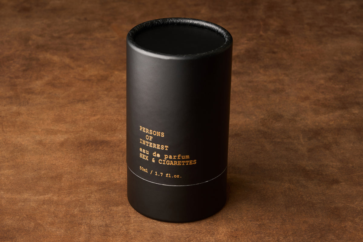 A black cylindrical bottle bearing gold lettering—Persons of Interest Sex & Cigarettes Eau de Parfum, 50 ml / 1.7 fl. oz.—rests on a brown textured surface, capturing the smoky allure and warm tobacco-bergamot notes of this distinctive fragrance.