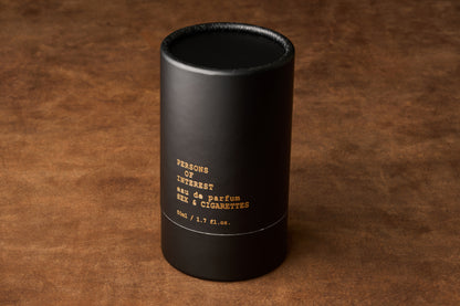 A black cylindrical bottle bearing gold lettering—Persons of Interest Sex & Cigarettes Eau de Parfum, 50 ml / 1.7 fl. oz.—rests on a brown textured surface, capturing the smoky allure and warm tobacco-bergamot notes of this distinctive fragrance.