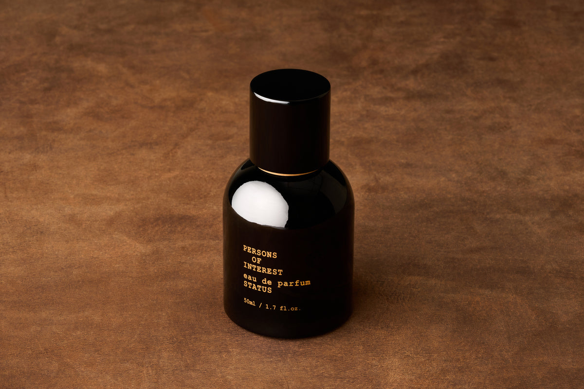 A black bottle of Persons of Interest Status Eau de Parfum, featuring notes of amber and sandalwood, sits on a brown textured surface. The label reads “eau de parfum STATUS 50mL / 1.7 fl. oz.”.