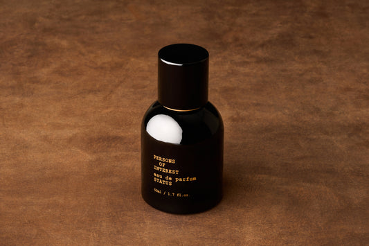 A black bottle of Persons of Interest Status Eau de Parfum, featuring notes of amber and sandalwood, sits on a brown textured surface. The label reads “eau de parfum STATUS 50mL / 1.7 fl. oz.”.