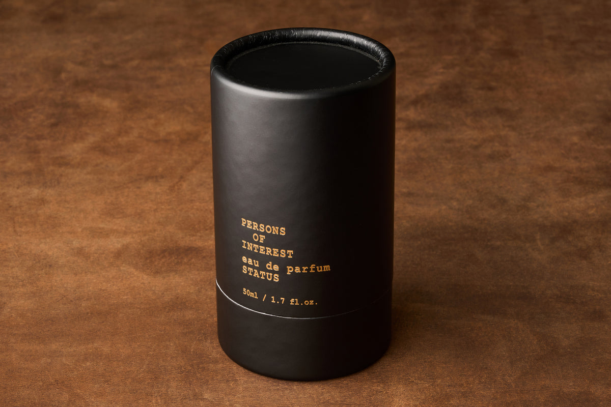 A black cylindrical box labeled “Persons of Interest Status Eau de Parfum, 50 ml / 1.7 fl. oz.” rests on a brown textured surface, suggesting the refined amber sandalwood fragrance crafted in Ontario, Canada.
