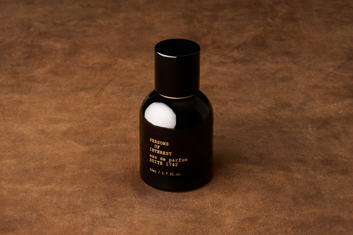 A black perfume bottle with gold text rests on a brown textured surface. The label reads PERSONS OF INTEREST Suite 1742 Eau de Parfum 50ML / 1.7 fl. oz., suggesting a traditional fragrance in the style of Crown Shaving Co, blending classic elegance with modern sophistication.