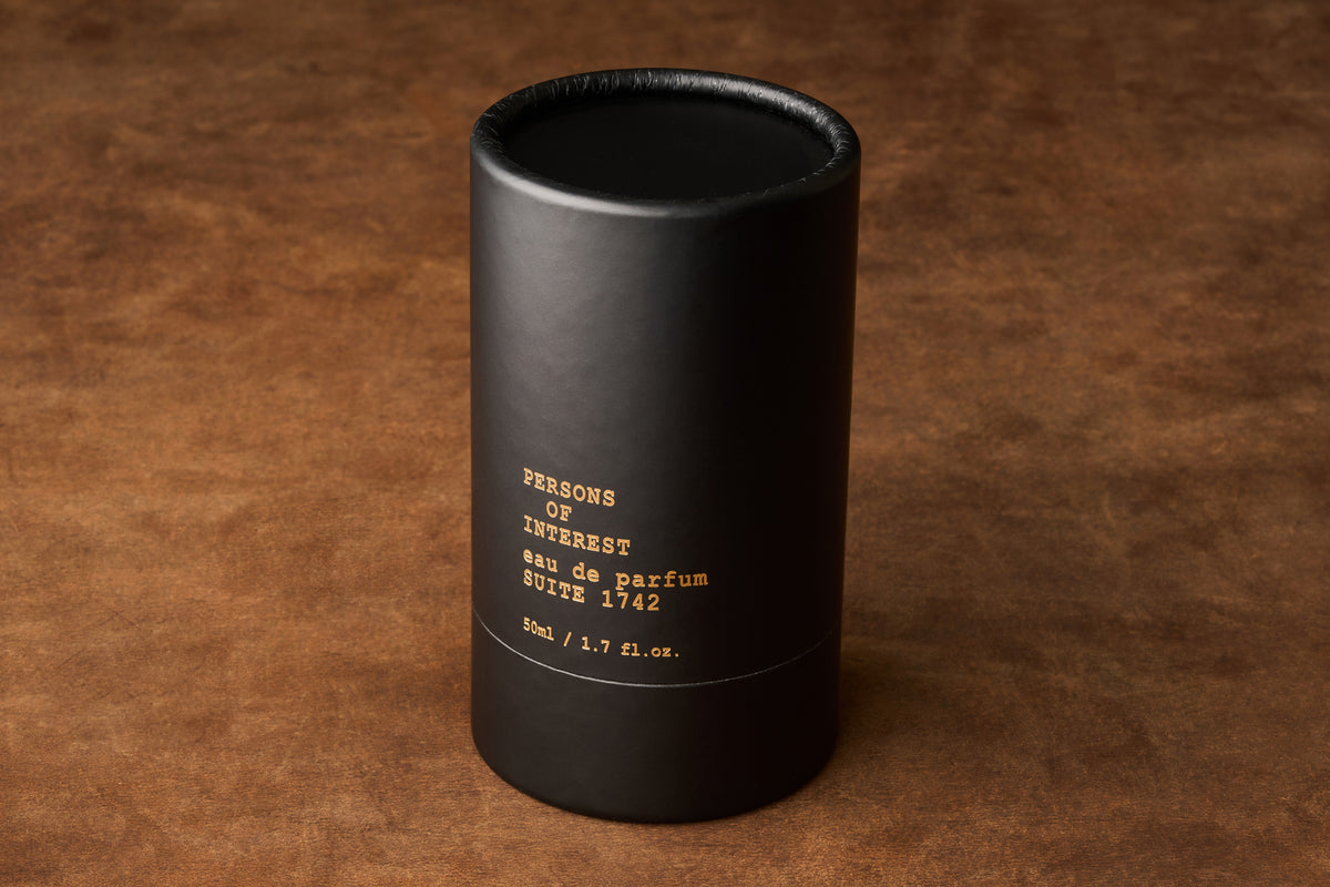 A black cylindrical container labeled Persons of Interest Suite 1742 Eau de Parfum, 50ml / 1.7 fl. oz., evoking a classic scent, rests on a brown textured surface.