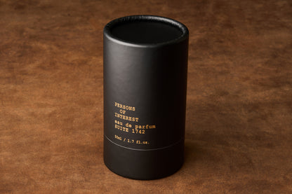 A black cylindrical container labeled Persons of Interest Suite 1742 Eau de Parfum, 50ml / 1.7 fl. oz., evoking a classic scent, rests on a brown textured surface.