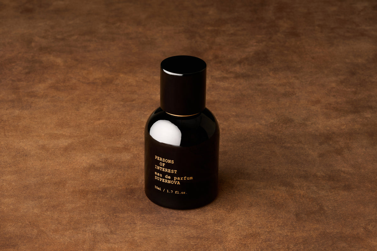 A black perfume bottle labeled Persons of Interest Supernova Eau de Parfum sits on a brown textured surface, casting a soft shadow. This sleek presentation perfectly matches the gender-neutral fragrance inside, which offers an intriguing and balanced citrus floral scent.