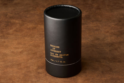 A black cylindrical box labeled “Persons of Interest Supernova Eau de Parfum” in a 50ML (1.7 fl. oz.) size sits upright on a brown textured surface, suggesting the luminous citrus floral notes of this unisex fragrance inside.