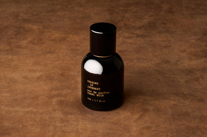 A black perfume bottle labeled Persons of Interest Tabac Noir stands upright on a textured brown surface, showcasing a refined unisex fragrance crafted in Ontario.