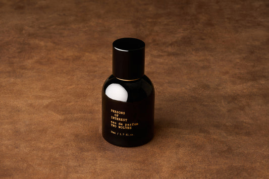 A black bottle of Persons of Interest Two Wolves Eau de Parfum rests on a brown, textured surface, suggesting the light fragrance inside—an intriguing blend of jasmine and vanilla.