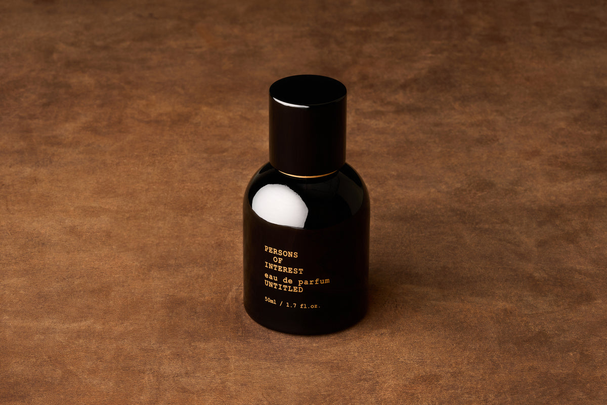 A black bottle with gold lettering sits atop a brown textured surface. Its label displays “PERSONS OF INTEREST, Eau de Parfum, UNTITLED, 50ml / 1.7 fl. oz.” This is the Persons of Interest Untitled Eau de Parfum—an intriguing, gender-neutral fragrance crafted in Ontario, Canada that captivates the senses with its mysterious allure.