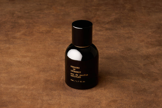 A black bottle with gold lettering sits atop a brown textured surface. Its label displays “PERSONS OF INTEREST, Eau de Parfum, UNTITLED, 50ml / 1.7 fl. oz.” This is the Persons of Interest Untitled Eau de Parfum—an intriguing, gender-neutral fragrance crafted in Ontario, Canada that captivates the senses with its mysterious allure.