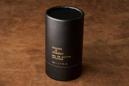 A black cylindrical box labeled “Persons of Interest Untitled Eau de Parfum 50ml / 1.7 fl. oz.” rests on a brown suede surface, alluding to the gender-neutral, paradoxical fragrance crafted in Ontario, Canada.