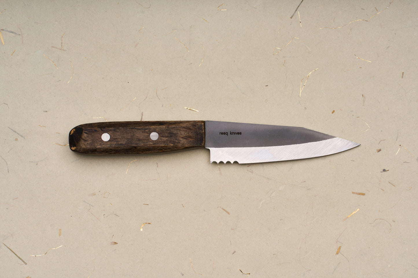 The RESQ Knives Korpi Camp Kitchen Knife, featuring a dark wooden handle and a short, sharp blade made from recycled stainless steel, rests on a beige surface flecked with scattered specks and fibers.