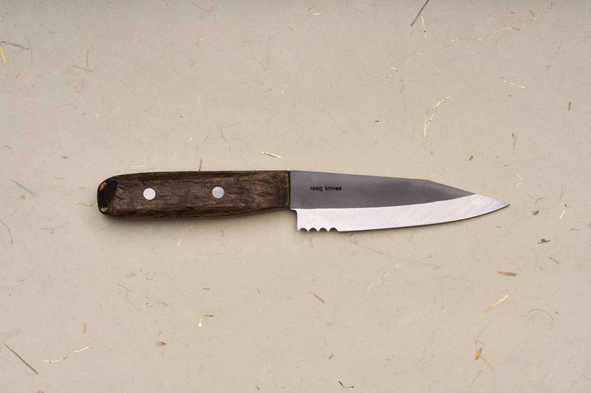 The RESQ Knives Korpi Camp Kitchen Knife, featuring a dark wooden handle and a short, sharp blade made from recycled stainless steel, rests on a beige surface flecked with scattered specks and fibers.