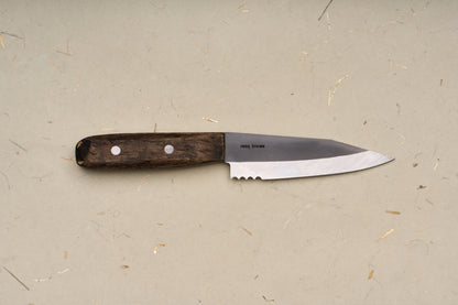 The RESQ Knives Korpi Camp Kitchen Knife, featuring a dark wooden handle and a short, sharp blade made from recycled stainless steel, rests on a beige surface flecked with scattered specks and fibers.