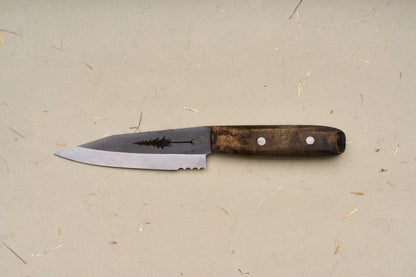 The RESQ Knives Korpi Camp Kitchen Knife, featuring a dark wooden handle with silver rivets and a partially serrated blade made from recycled stainless steel adorned with a tree silhouette design, rests on a beige background subtly flecked with texture.