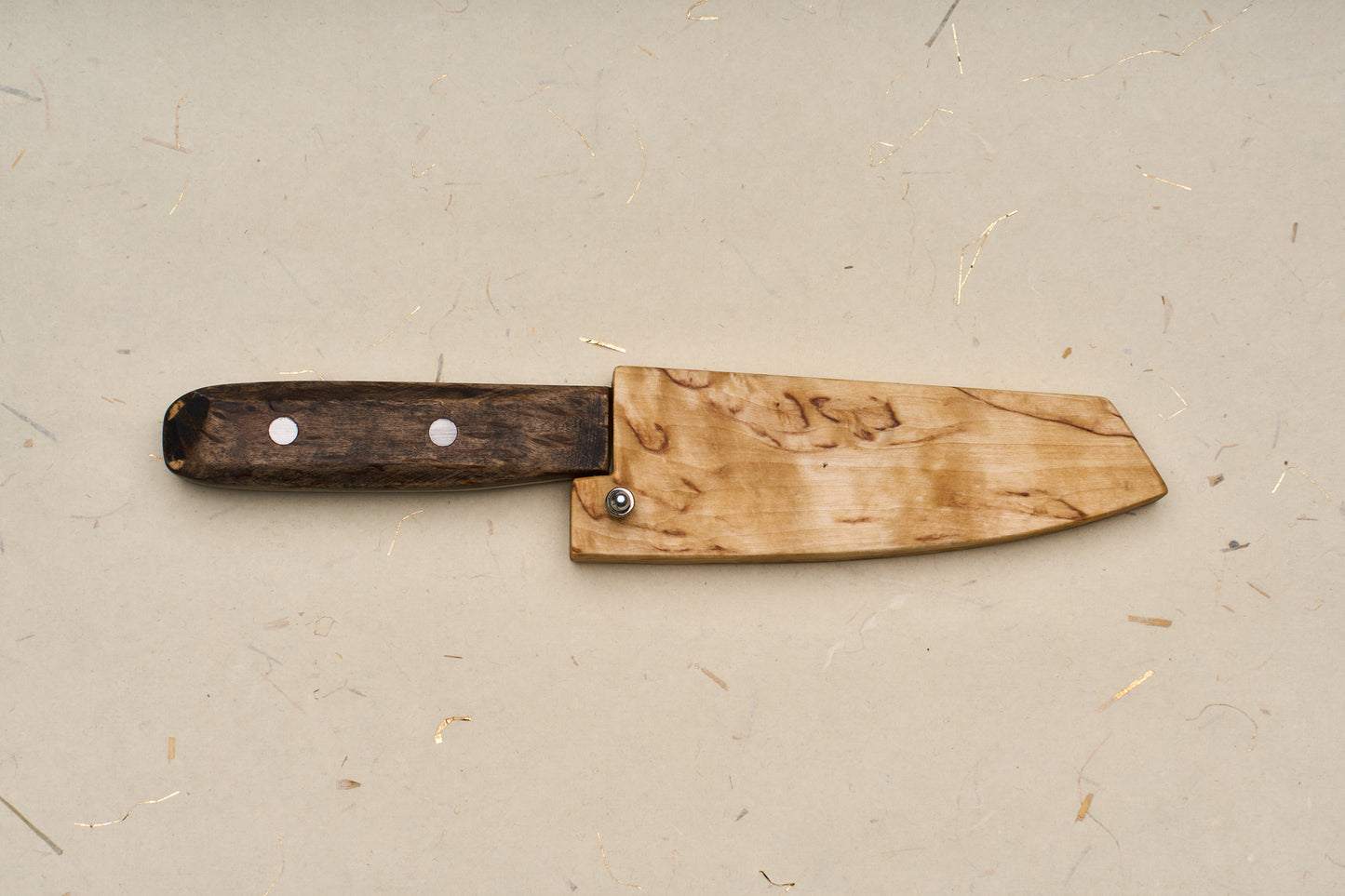 The RESQ Knives Korpi Camp Kitchen Knife, featuring a dark wooden handle and a light wooden sheath, rests on a beige surface with subtle flecks and fibers. Its blade is expertly crafted from recycled stainless steel.