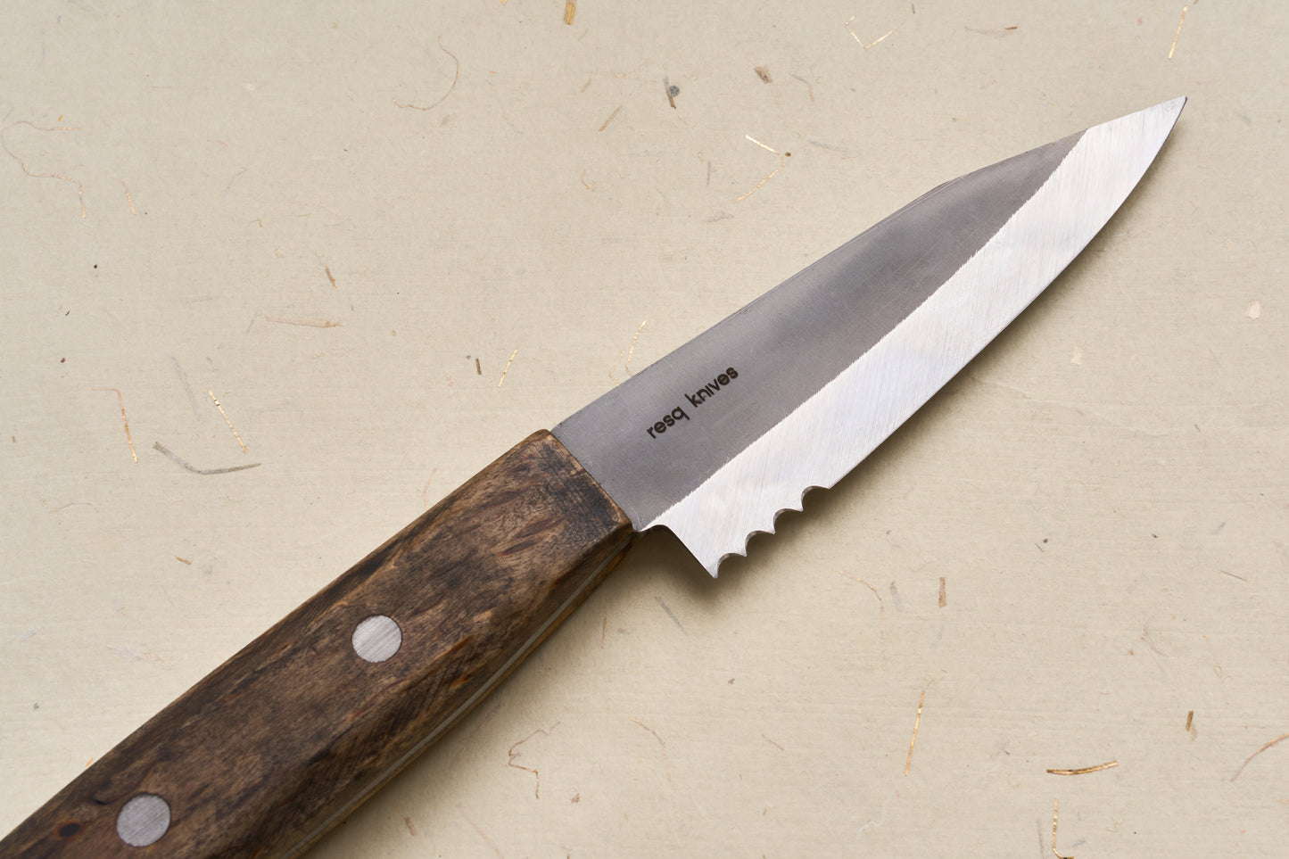 A RESQ Knives Korpi Camp Kitchen Knife, featuring a wooden handle and a partially serrated blade made from recycled stainless steel, is lying on a beige surface.