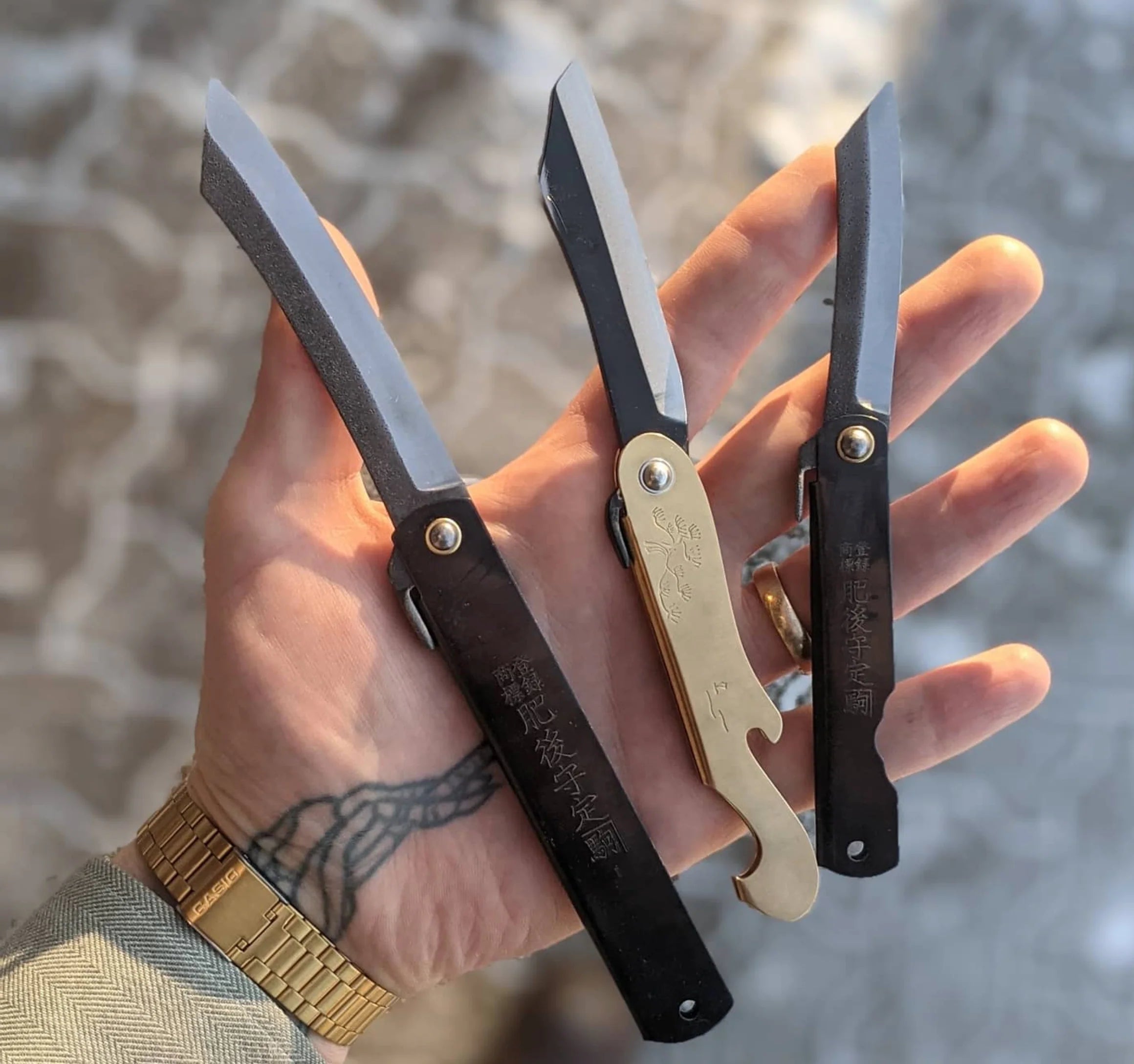 Three folding knives held in a hand with a blurred natural background