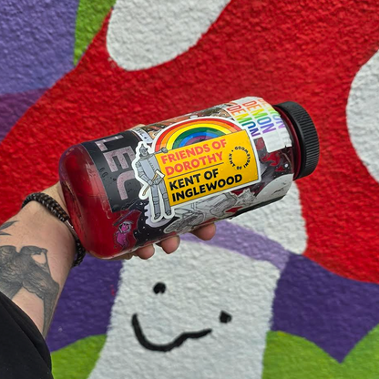 A tattooed arm holds a sticker-covered bottle—proudly displaying the vibrant Kent of Inglewood Pride Sticker—in front of a colorful, abstract mural with a smiling face and bold swathes of red, green, purple, and white.
