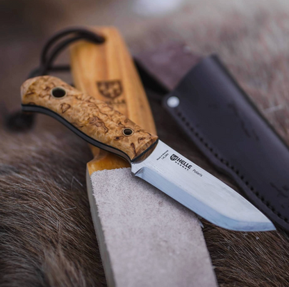 A Helle Knives Polaris fixed-blade knife with a wooden handle rests on a sharpening stone, accompanied by a leather sheath and wooden board in the background, all arranged on a fur surface—an excellent example of quality outdoor knives.