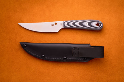 A Spyderco Bow River knife with a patterned handle rests atop a black leather sheath on an orange background.