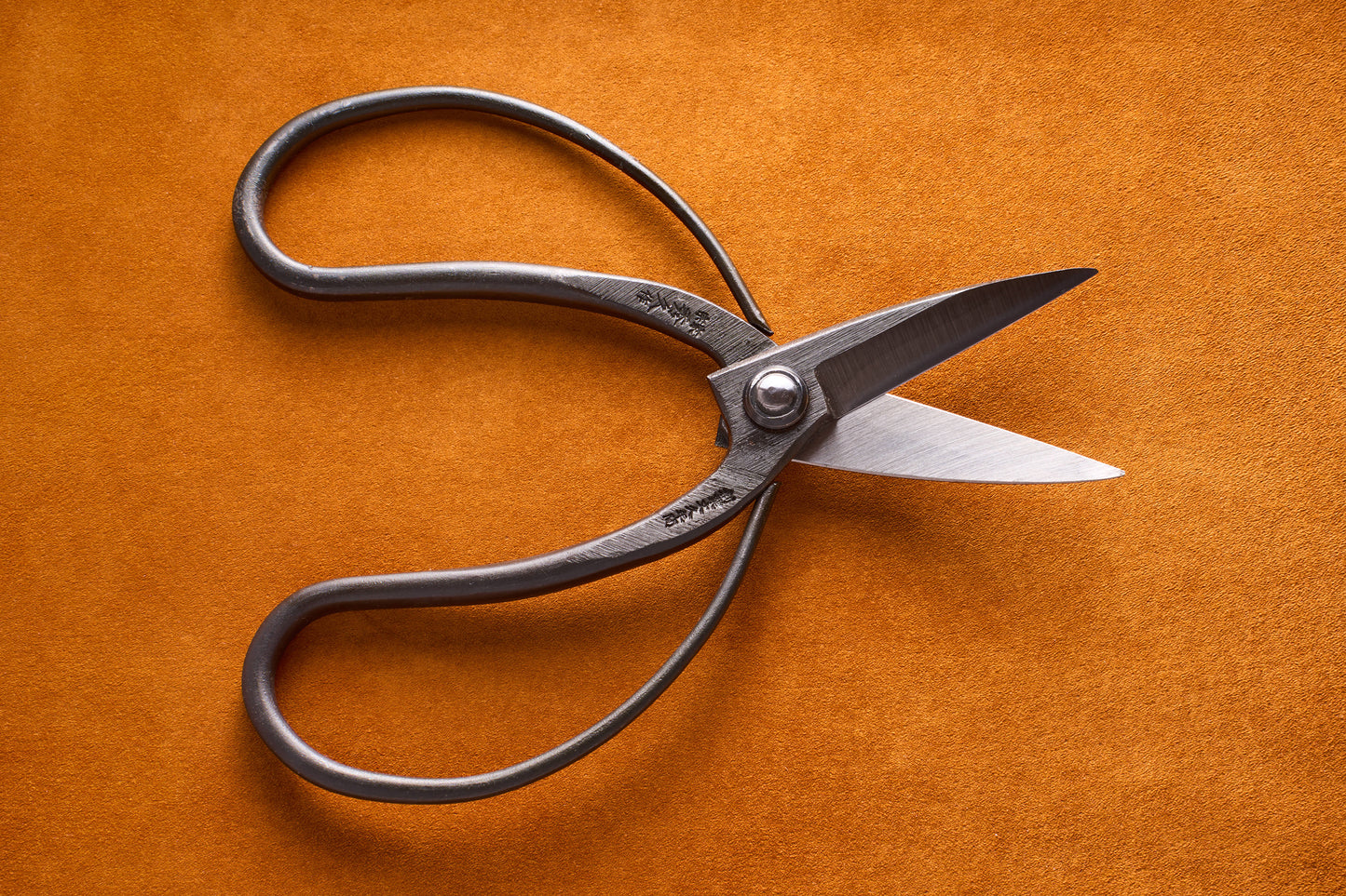 The Toyama Okubo Black Bonsai Shears 200mm T1, featuring curved handles and sharp blades reminiscent of vintage metal scissors, are placed on an orange textured surface and photographed from above.