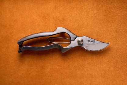 The Toyama Pruning Offset Shears 200mm T13, featuring a curved blade and metal spring, rest on an orange textured surface, embodying the precision and artistry of traditional Japanese gardening.