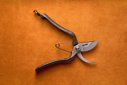 A pair of Toyama Pruning Shears 200mm T14 with a curved blade and spring mechanism lies open on an orange textured surface, exemplifying the precision tools characteristic of a Japanese garden.