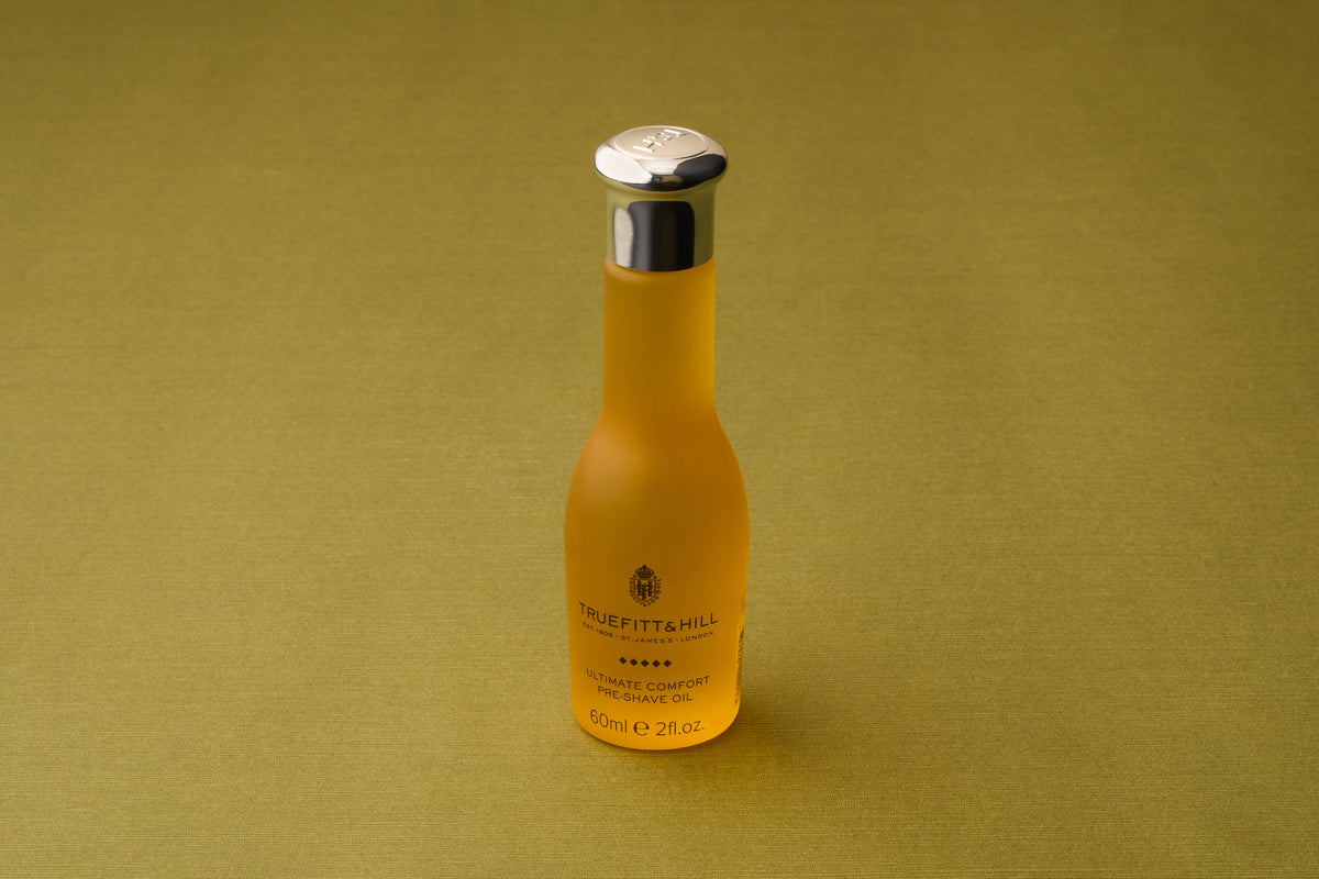 A frosted glass bottle featuring a silver cap and bearing the Truefitt & Hill Ultimate Comfort Pre-Shave Oil name (60ml), renowned as a premium men’s grooming essential, rests atop a green textured surface that recalls the refined luxury of classic Victorian shaving traditions.