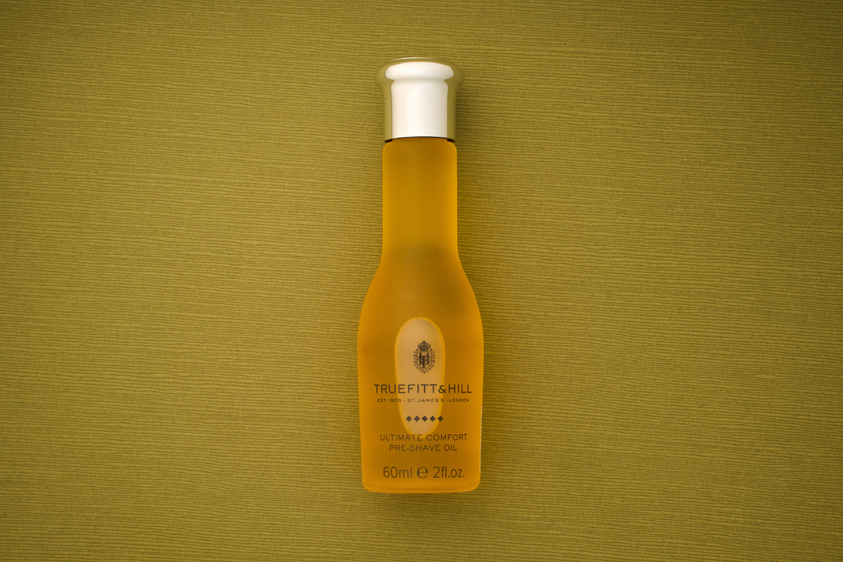 A bottle of Truefitt & Hill Ultimate Comfort Pre-Shave Oil, 60ml (2 fl. oz.), featuring a white cap, rests on a textured olive-green background, embodying the timeless sophistication of Victorian-era shaving traditions in contemporary men’s grooming.