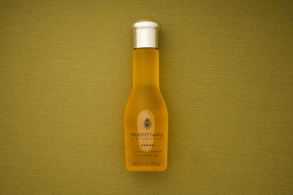 A bottle of Truefitt & Hill Ultimate Comfort Pre-Shave Oil, 60ml (2 fl. oz.), featuring a white cap, rests on a textured olive-green background, embodying the timeless sophistication of Victorian-era shaving traditions in contemporary men’s grooming.
