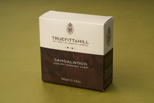 A box of Truefitt & Hill Sandalwood Luxury Shaving Soap rests on a green surface. The beige and brown packaging is adorned with gold accents and text, indicating a net weight of 100g (3.5oz). Designed for those who value classic shaving rituals, this sandalwood luxury shaving soap delivers an indulgent grooming experience.
