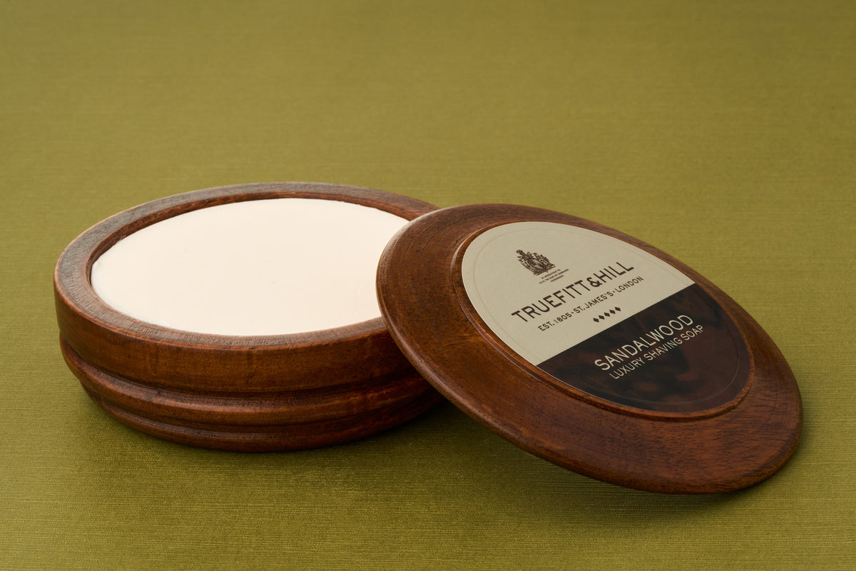 A round wooden container of Truefitt & Hill Sandalwood Luxury Shaving Soap, its lid partially open to reveal the white soap inside, rests on a green surface—a classic choice for those who appreciate traditional shaving.