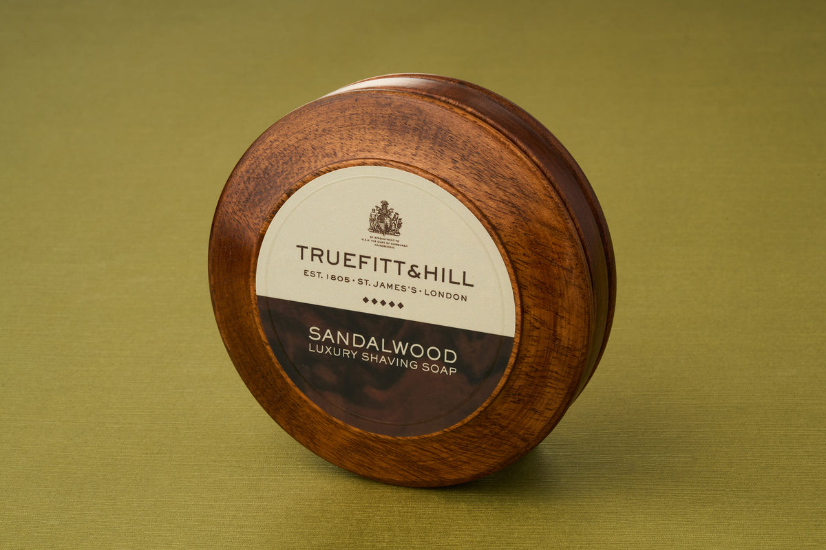 A round wooden container of Truefitt & Hill Sandalwood Luxury Shaving Soap rests atop an olive green surface, its label displayed—an enduring must-have for enthusiasts of classic wet shaving and the warm, refined scent of sandalwood.