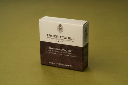 A box of Truefitt & Hill Sandalwood Luxury Shaving Soap refill stands upright on a green-toned surface. Its cream and brown packaging—crafted to pair with a refillable wooden shaving bowl—features classic product details and weight, exuding timeless elegance.