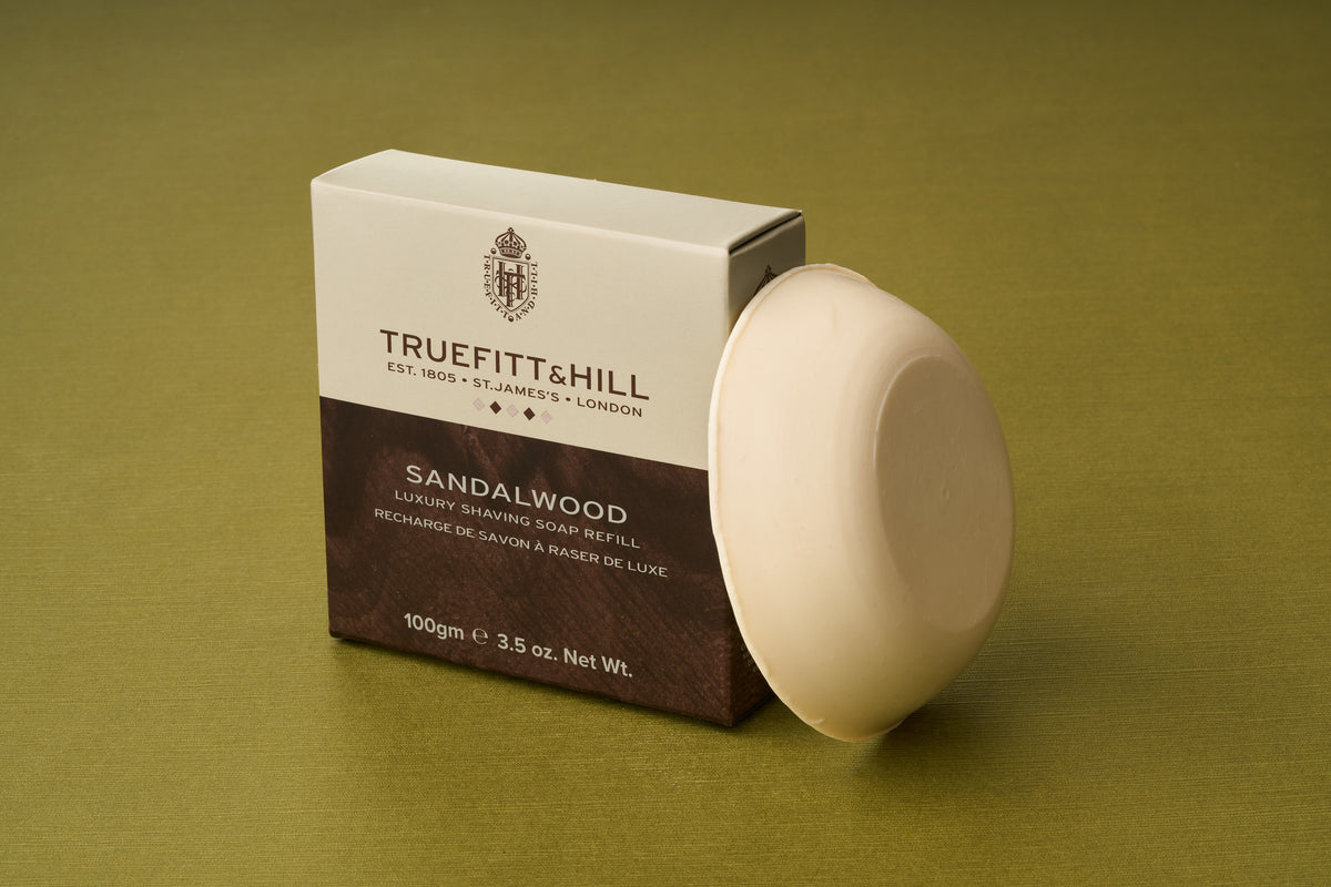 A round bar of Truefitt & Hill Sandalwood Luxury Shaving Soap is pictured resting against a Truefitt & Hill branded box on a green surface. The box displays product details and weight, and both items are well-lit. This luxurious shaving soap can be used as a refill in your favorite refillable wooden shaving bowl, offering a blend of classic tradition and modern convenience.