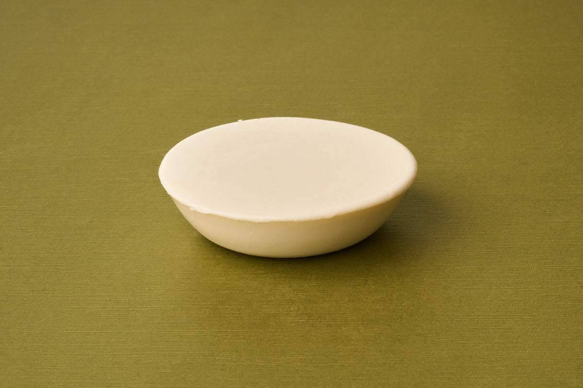 A round, white bar of Truefitt & Hill Sandalwood Luxury Shaving Soap rests on a smooth olive green surface against a simple backdrop, evoking the enduring elegance of traditional shaving.