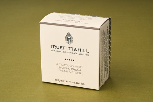 A beige box of Truefitt & Hill Ultimate Comfort Shave Cream, renowned for its Victorian shave tradition, sits on a green surface, featuring black text and a crest logo on the front.