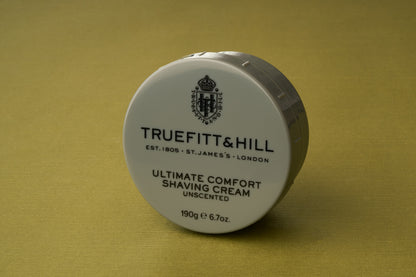 A round white container of Truefitt & Hill Ultimate Comfort Shave Cream rests on a gold surface. Designed for those who appreciate the timeless elegance of a Victorian shave, the label specifies it is unscented and contains 190g (6.7oz).