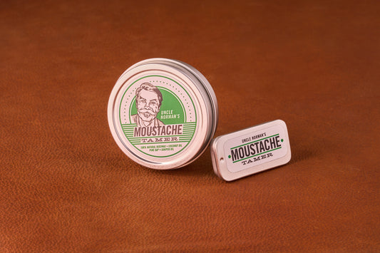 Two tins of Uncle Norman’s Moustache Tamer, a beeswax-based styling balm, highlighting versatility and hold. Features a round and a travel-sized rectangular tin.